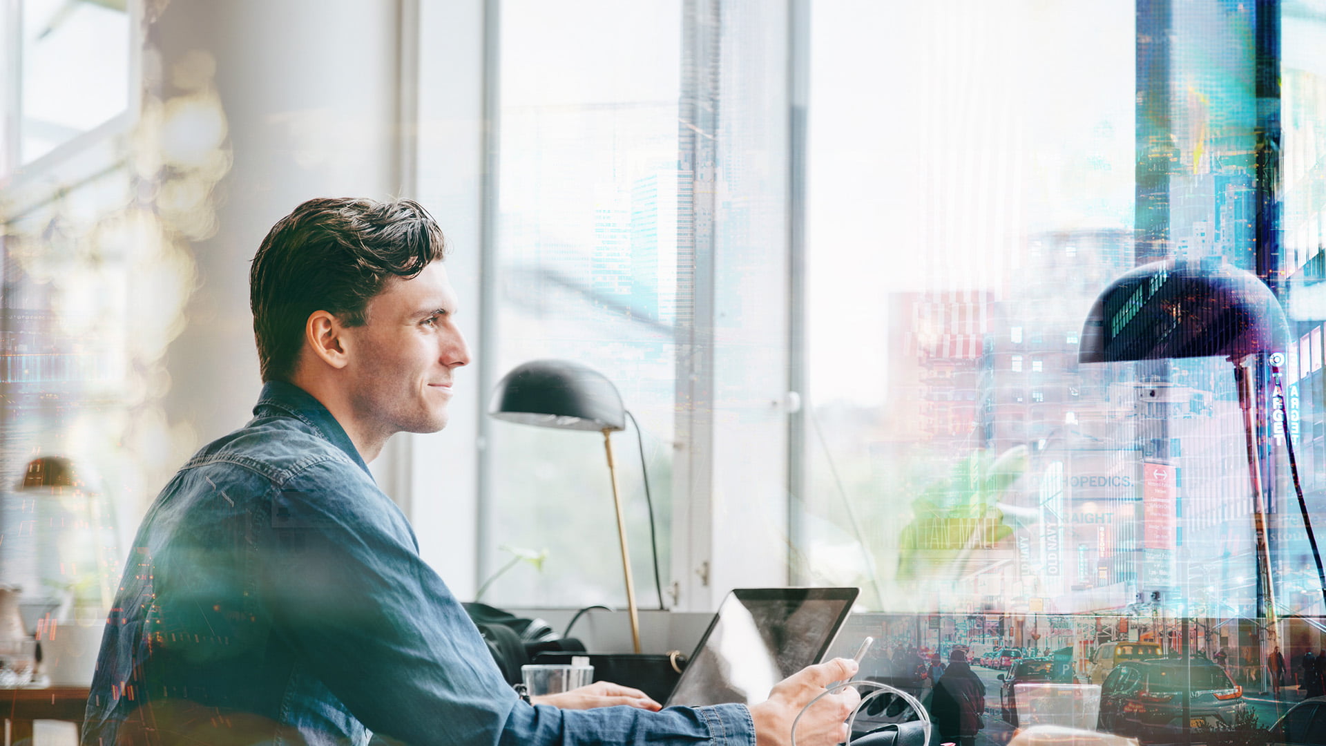 Man i jeansskjorta som sitter på ett café med laptop framför sig och blickar ut genom ett fönster