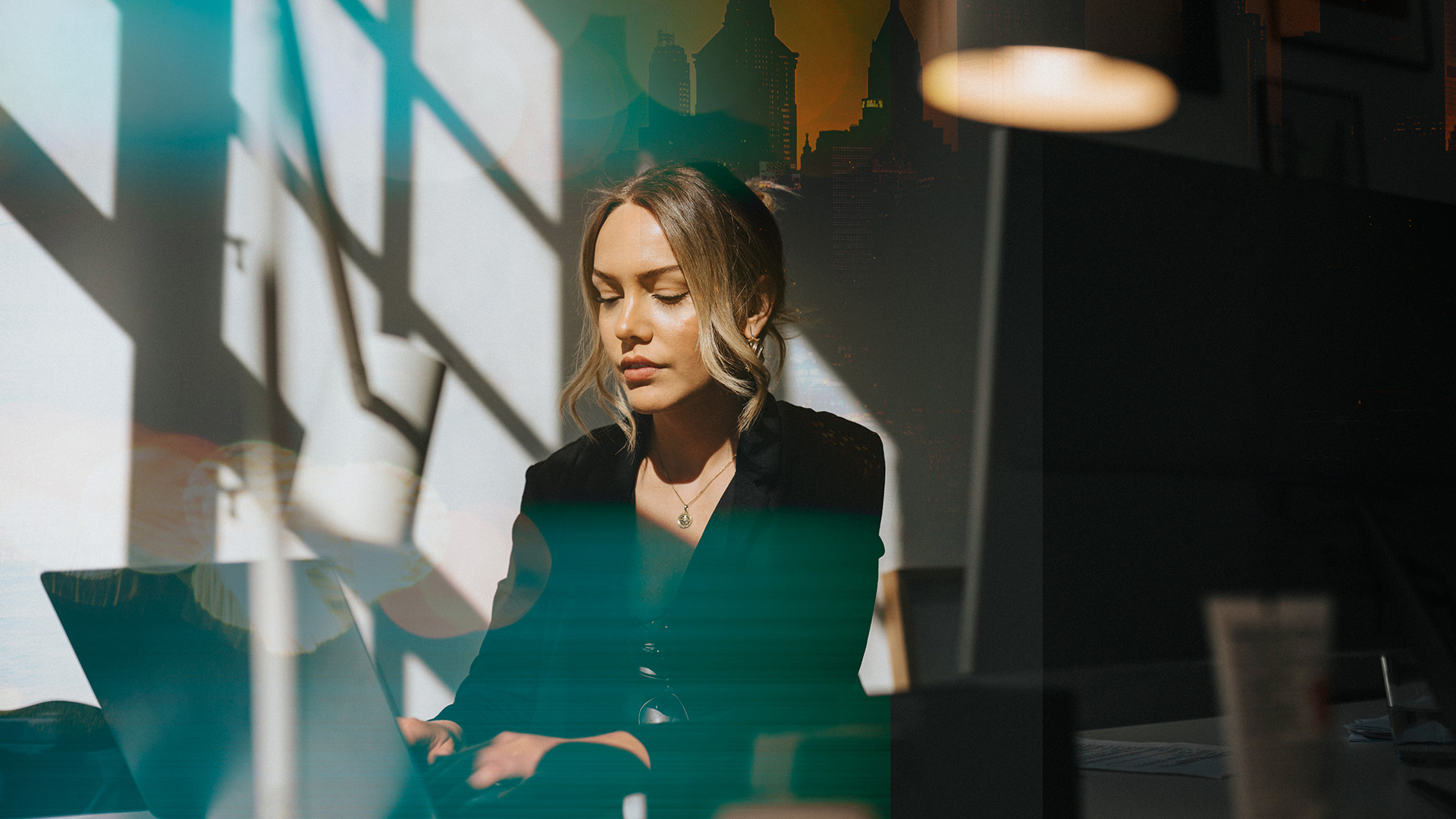 En kvinna sitter och jobbar vid sin laptop. På väggen bakom ser man skuggorna av ett fönster.