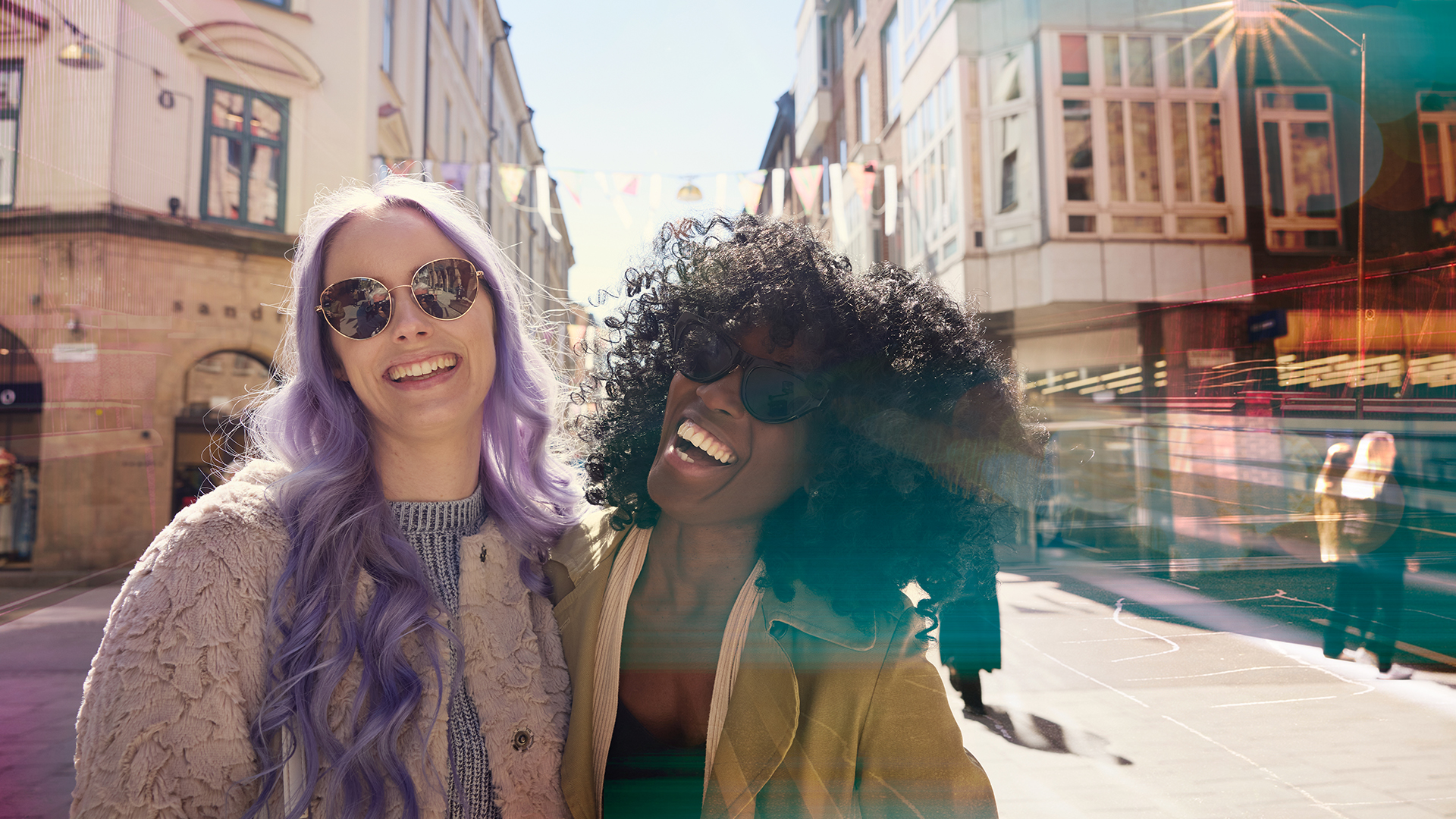 Två kvinnor med solglasögon med en gatuvy i bakgrunden som skrattar mot kameran